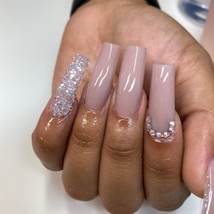 May include: Close-up of a hand with long, nude-colored acrylic nails. The nails are decorated with rhinestones, with one nail featuring a full row of rhinestones.
