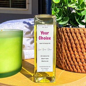 May include: A rectangular glass bottle of "Your Choice" bath and body oil with a black cap. The label includes "Hydrate, Soften, Repair". The bottle is on a wooden tray with a green candle and a woven basket in the background. The text "Essential Spa Company" is at the bottom.