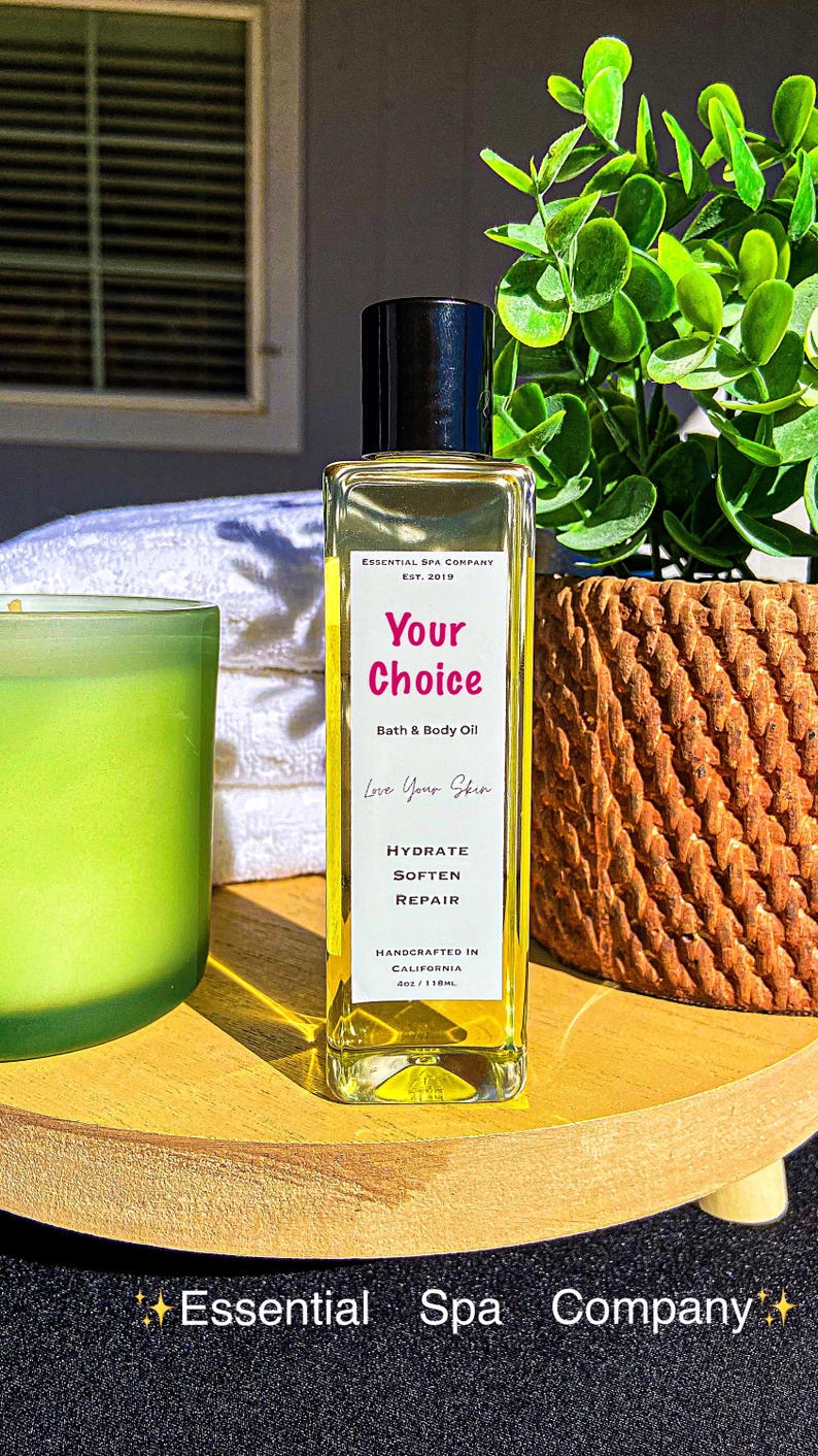 May include: A rectangular glass bottle of "Your Choice" bath and body oil with a black cap. The label includes "Hydrate, Soften, Repair". The bottle is on a wooden tray with a green candle and a woven basket in the background. The text "Essential Spa Company" is at the bottom.