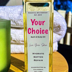 May include: A rectangular glass bottle of bath and body oil with a black cap. The label reads "Your Choice" in pink, with "Bath & Body Oil" below. The bottle is 4oz/118mL and says "Handcrafted in California."