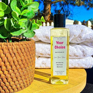 May include: A clear rectangular bottle of bath and body oil with a black cap, labeled "Your Choice." The bottle sits on a wooden tray with a woven basket and a plant. White towels are in the background.