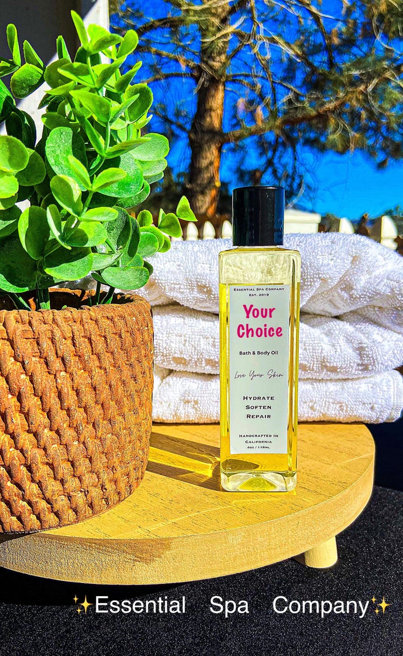 May include: A clear rectangular bottle of bath and body oil with a black cap, labeled "Your Choice." The bottle sits on a wooden tray with a woven basket and a plant. White towels are in the background.
