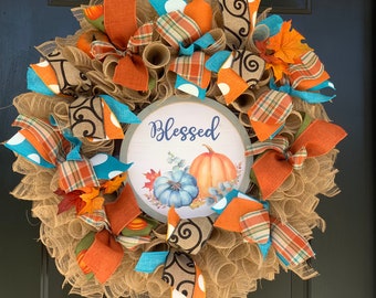 May include: A burlap wreath decorated with orange, blue, and brown ribbons, fall leaves, and a sign that reads "Blessed" with a pumpkin and floral design.