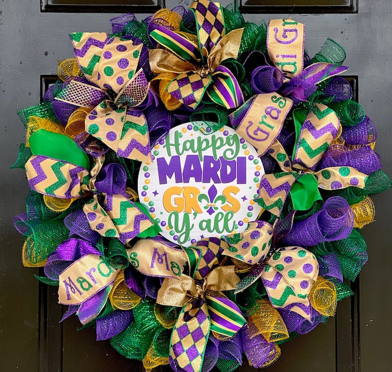 Op de afbeelding: Een feestelijke Mardi Gras-krans met groen, paars en goudkleurig gaas. De krans heeft decoratieve linten, strikken en een centraal bord met de tekst "Happy Mardi Gras Y'all". Het bord heeft een fleur-de-lis-symbool.