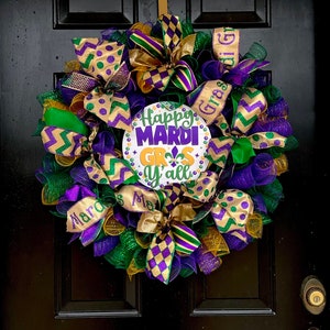 Op de afbeelding: Een feestelijke Mardi Gras-krans met paarse, groene en gouden linten en gaas. De krans heeft een centraal bord met de tekst "Happy Mardi Gras y'all" met een fleur-de-lis-symbool. Het hangt aan een zwarte deur.