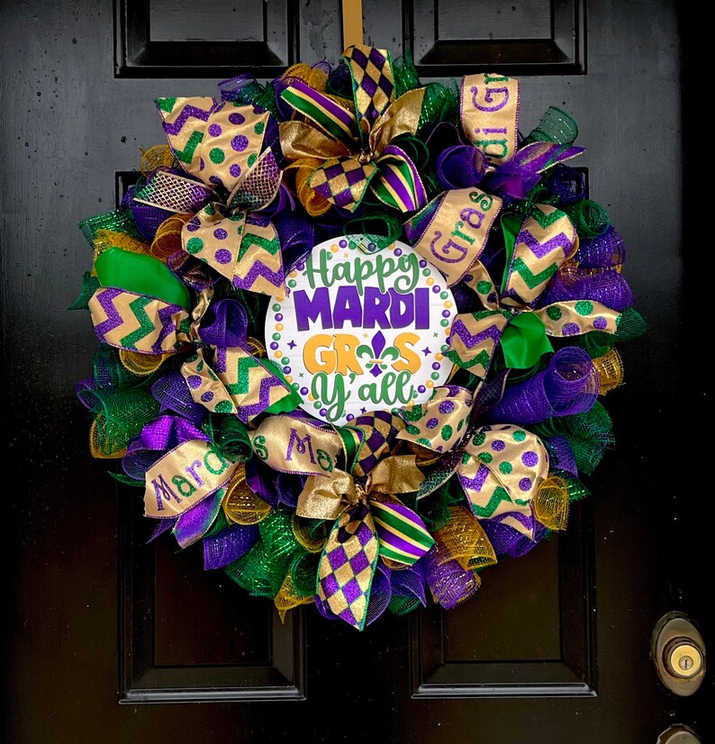Op de afbeelding: Een feestelijke Mardi Gras-krans met paarse, groene en gouden linten en gaas. De krans heeft een centraal bord met de tekst "Happy Mardi Gras y'all" met een fleur-de-lis-symbool. Het hangt aan een zwarte deur.