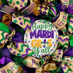 Op de afbeelding: Een feestelijke Mardi Gras-krans met paarse, groene en gouden linten en gaas. De krans heeft een centraal bord met de tekst "Happy Mardi Gras Y'all" in paars, groen en goud. De krans is versierd met strikken en decoratieve elementen.