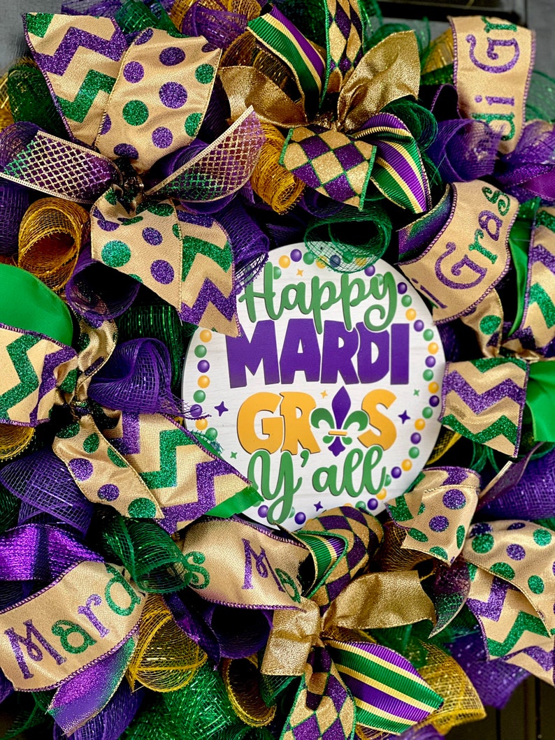 Op de afbeelding: Een feestelijke Mardi Gras-krans met paarse, groene en gouden linten en gaas. De krans heeft een centraal bord met de tekst "Happy Mardi Gras Y'all" in paars, groen en goud. De krans is versierd met strikken en decoratieve elementen.