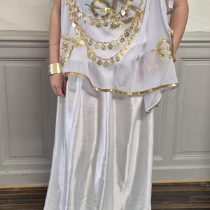 May include: White costume with gold detailing, comprising a draped top embellished with gold coins and trim, and a long, flowing skirt. The outfit includes gold jewellery and a cuff bracelet, evoking a classical theme.