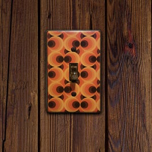 May include: A retro-style light switch with a decorative cover featuring a repeating pattern of orange, brown, and black circles. The switch is mounted on a wooden wall with vertical planks.