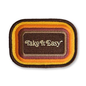 May include: A rectangular patch with rounded corners, featuring a retro color scheme of brown, orange, yellow, and purple. The patch has the words "Take It Easy" embroidered in a cursive font, perfect for jackets, bags, or other fabric items.