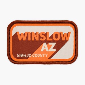 May include: A rectangular embroidered patch with a dark brown border. The design features the orange text WINSLOW AZ and white text NAVAJO COUNTY against a background of diagonal orange and off-white sections with a dark brown geometric shape.