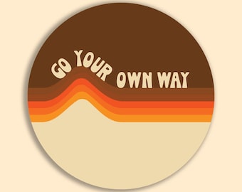 May include: A round sticker with a retro design. The top half is brown, with the words "GO YOUR OWN WAY" in white. Below are orange and yellow horizontal stripes. The bottom half is beige.