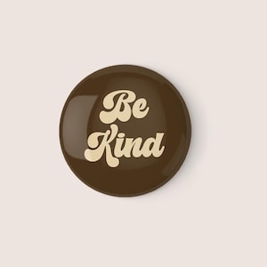 May include: A brown round button with the text "Be Kind" in a retro font in cream color.