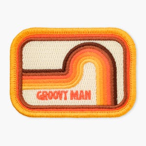 May include: A rectangular embroidered patch with rounded corners, featuring a cream-colored background and concentric stripes in shades of brown, orange, and yellow. The text GROOVY MAN is stitched in orange letters at the bottom center.