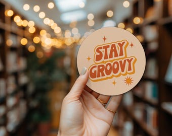 May include: A round, beige coaster with the words "STAY GROOVY" in orange and yellow lettering. The coaster has a retro design with star and sunburst accents. The coaster is held in a hand.