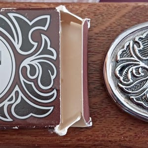 May include: A silver compact mirror with a Western saddle design. The compact has a brown box with the text "Avon Western Saddle Belt Buckle".