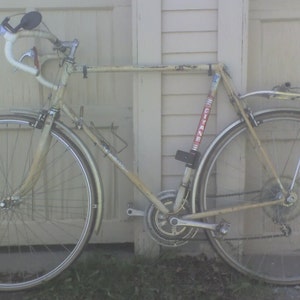 May include: A white vintage road bike with a silver frame and silver wheels. The bike has a silver handlebar with a black mirror, a silver seat, and a silver chain. The bike has a black and red sticker that says "Gitane" on the frame.