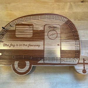 May include: A wooden cribbage board shaped like a camper van, with a brown and white wood grain finish. The board has a hole pattern for pegs and the text "The Joy is in the Journey" is engraved on the front. The board also has a space for a name to be engraved.