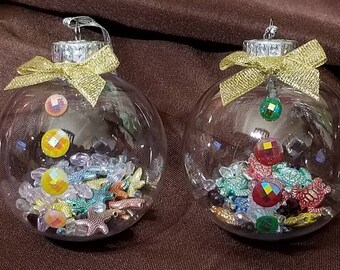 May include: Two clear glass Christmas ornaments with gold bows and various colorful beads and sea-themed charms inside. The ornaments are filled with small, iridescent beads, starfish, and sea turtles.