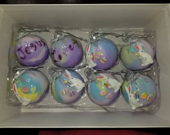 May include: Eight Easter-themed glass ornaments with a pastel purple, blue, and yellow color scheme. Each ornament features a white bunny with a different Easter-themed accessory. Two of the ornaments have personalized names, "Jenny" and "Love", written in purple glitter.
