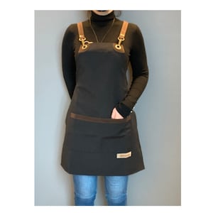 Peut inclure: Un tablier noir avec des bretelles en cuir marron et une poche en cuir marron. Le tablier a une boucle en laiton et un petit patch en cuir marron avec le mot "Remarkable" dessus.