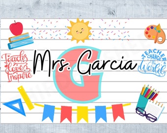 May include: Teacher desk decor featuring a large pink and blue letter G, a sun, a red apple, a stack of books, a ruler, a string of flags, a paint palette, a pencil holder with coloured pencils, glasses, and a notebook. The text "Mrs. Garcia" is written in black cursive. The text "Teacher Desk Decor 38X24" is written in black.