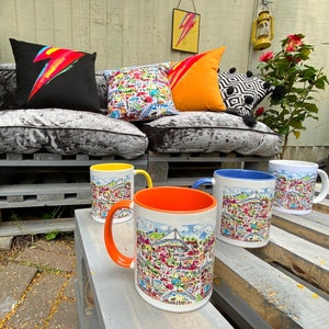 May include: Four ceramic mugs with colorful cityscapes printed on them. The mugs are sitting on a wooden surface. The mugs are yellow, blue, orange, and white.