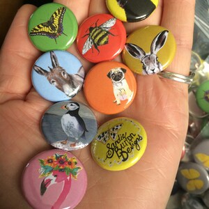 May include: A hand holding a variety of colourful round buttons with different animal and insect designs. The buttons feature a bee, a butterfly, a cat, a donkey, a rabbit, a pug, a puffin, and a flamingo. One button has the text "Sandie Button Designs".