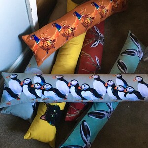 May include: A collection of colorful, decorative bolster pillows with various animal and nature-themed designs. The pillows feature images of puffins, a black cat, a pheasant, and a rabbit.