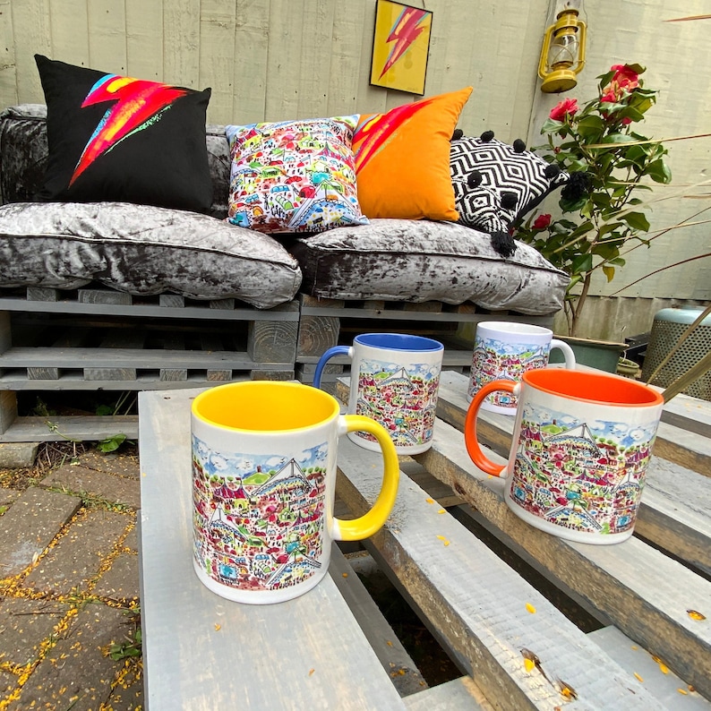 May include: Four ceramic mugs with colourful illustrations of a city skyline. The mugs are on a wooden surface with a light brown finish. Two of the mugs are yellow, one is blue, and one is orange.