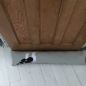 May include: A gray doorstop with a print of two seagulls. The doorstop is lying on a white wooden floor.