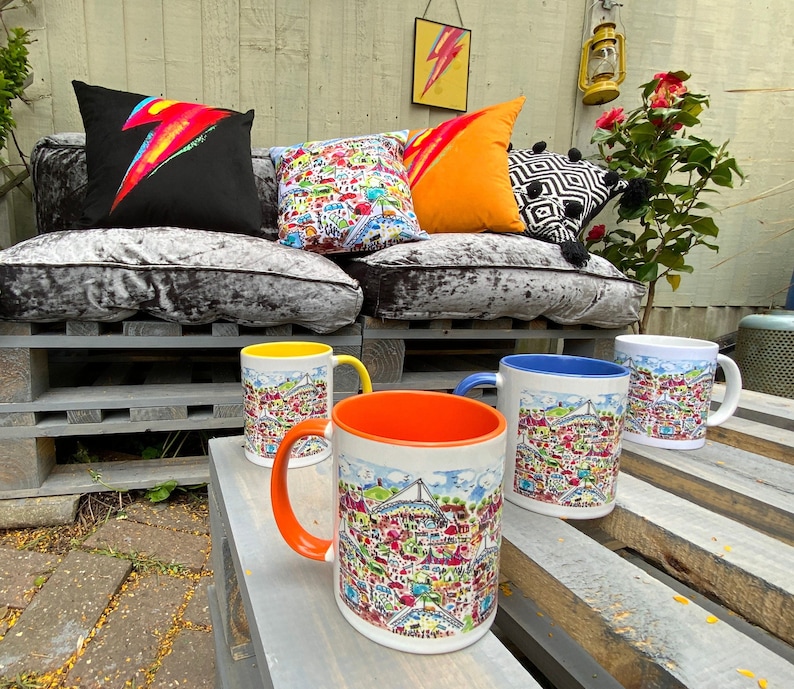 May include: Four ceramic mugs with colorful cityscapes printed on them. The mugs are sitting on a wooden surface. The mugs are yellow, blue, orange, and white.
