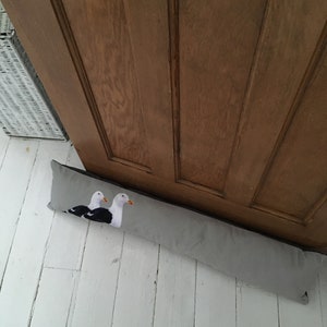May include: A gray doorstop with two embroidered seagulls on a white background. The doorstop is placed on a white wooden floor.