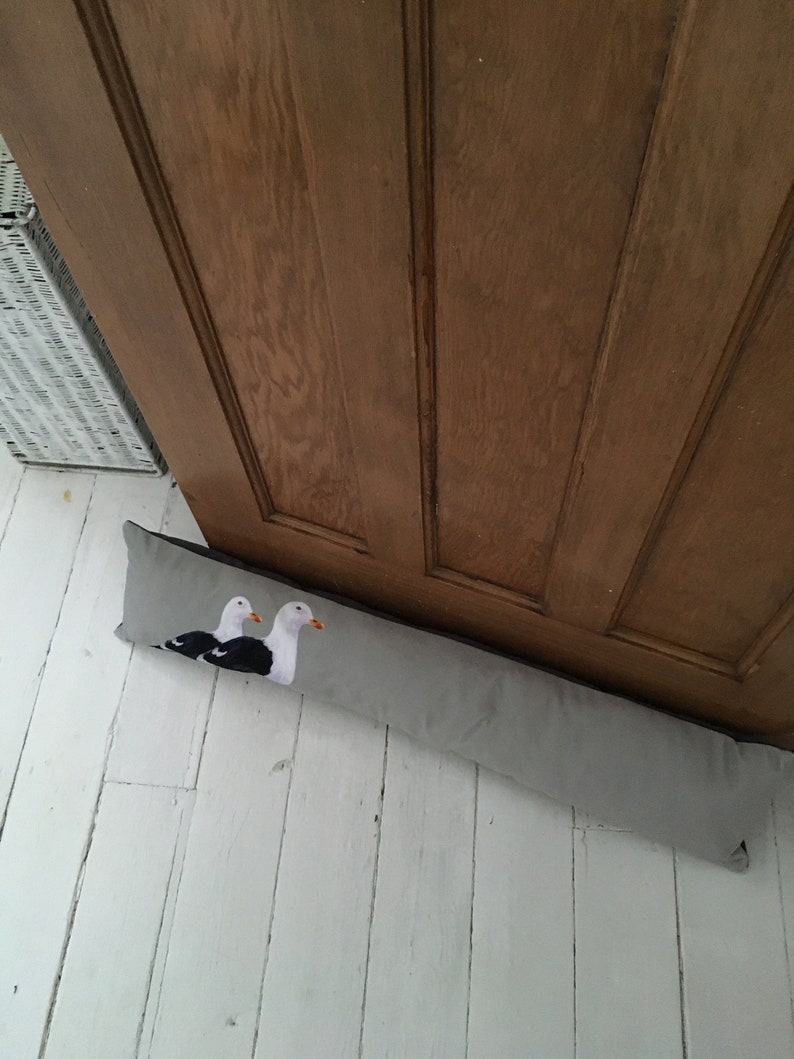 May include: A gray doorstop with two embroidered seagulls on a white background. The doorstop is placed on a white wooden floor.