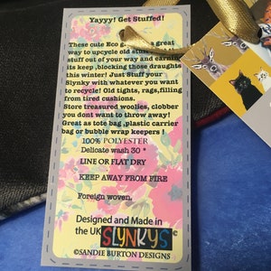 May include: A yellow and gray tag with the text "Yayyy! Get Stuffed!" and "These cute Eco... a great way to upcycle old stuff... its keep, blocking those draughts this winter! Just Stuff your Slynky with whatever you want to recycle! Old tights, rags, filling from tired cushions. Store treasured woolies, clobber you dont want to throw away! Great as tote bag, plastic carrier bag or bubble wrap keepers! 100% POLYESTER Delicate wash 30 * LINE OR FLAT DRY KEEP AWAY FROM FIRE Foreign woven. Designed and Made in the UK SLYNKYS OSANDIE BURTON DESIGNS"  The tag is attached to a blue and black striped fabric.