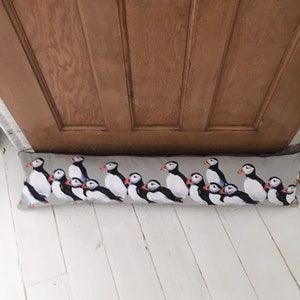 May include: A grey doorstop with a pattern of multiple puffins in black, white and orange. The doorstop is lying on a white wooden floor.