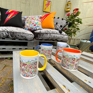 May include: Four ceramic mugs with colorful illustrations and handles. The mugs are on a wooden table with a light wood finish. The mugs are arranged in a row, with the yellow mug in the front.
