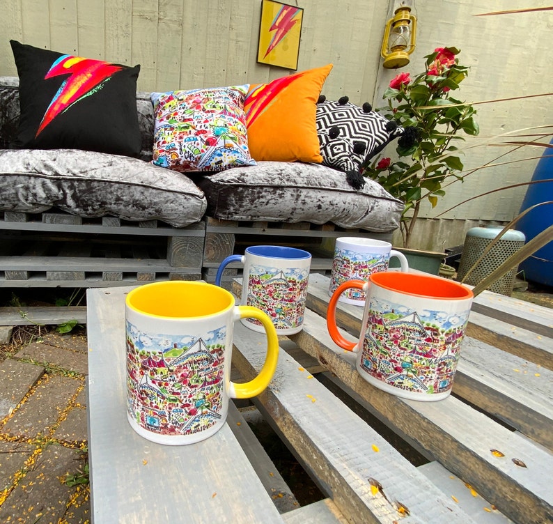 May include: Four ceramic mugs with colorful illustrations and handles. The mugs are on a wooden table with a light wood finish. The mugs are arranged in a row, with the yellow mug in the front.