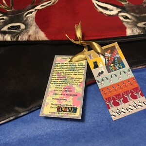 May include: A black fabric bag with a red and white deer print. The bag has a gold ribbon tied around it and a tag attached. The tag is white with red, orange, and blue illustrations of animals and the text "Yayyy! Get that stuff out of your way and earning its keep, blocking those draughts this winter! Just Stuff your Slynky with whatever you want to recycle! Old tights, rags, filling from tired cushions. Store treasured woolies, clobber you don't want to throw away! Great as tote bag, plastic carrier bag or bubble wrap keepers! 100% POLYESTER Delicate wash 30. LINE OR FLAT DRY KEEP AWAY FROM FIRE Foreign woven. Designed and Made in the UK SLYNKYS CSANDIE BURTON DESIGNS SLYNKYS".