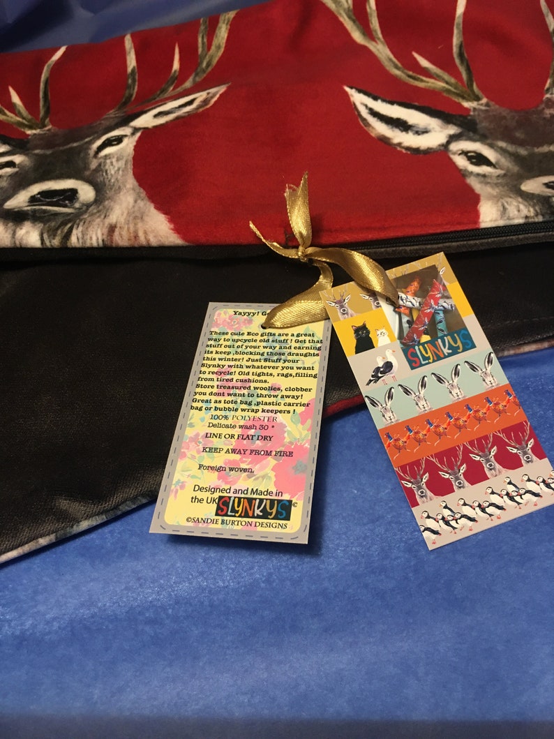 May include: A black fabric bag with a red and white deer print. The bag has a gold ribbon tied around it and a tag attached. The tag is white with red, orange, and blue illustrations of animals and the text "Yayyy! Get that stuff out of your way and earning its keep, blocking those draughts this winter! Just Stuff your Slynky with whatever you want to recycle! Old tights, rags, filling from tired cushions. Store treasured woolies, clobber you don't want to throw away! Great as tote bag, plastic carrier bag or bubble wrap keepers! 100% POLYESTER Delicate wash 30. LINE OR FLAT DRY KEEP AWAY FROM FIRE Foreign woven. Designed and Made in the UK SLYNKYS CSANDIE BURTON DESIGNS SLYNKYS".
