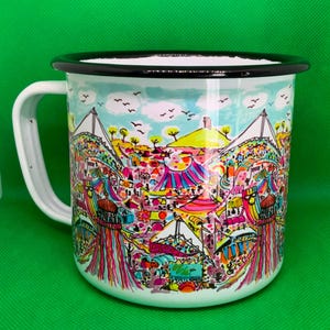 May include: White enamel mug with a black rim and handle. The mug features a colourful illustration of a festival scene with tents, trees, and people. The sky is light blue with birds.