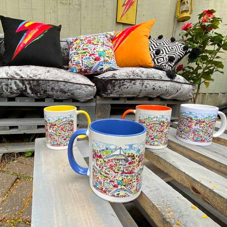 May include: Four ceramic mugs with colorful illustrations of a city skyline. The mugs are on a wooden surface with a light brown finish. The mugs are in different colors: yellow, blue, orange, and white.