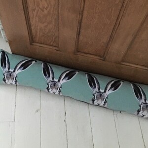 May include: A light blue doorstop with a repeating pattern of brown rabbit faces. The doorstop is lying on a white wooden floor.