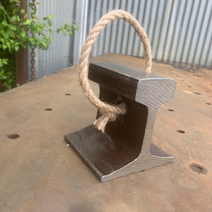 May include: A dark gray, industrial-style paperweight made from a section of railroad track. A loop of thick, natural-colored rope is attached to the top. The paperweight sits on a weathered metal surface, suggesting a rustic or vintage aesthetic.