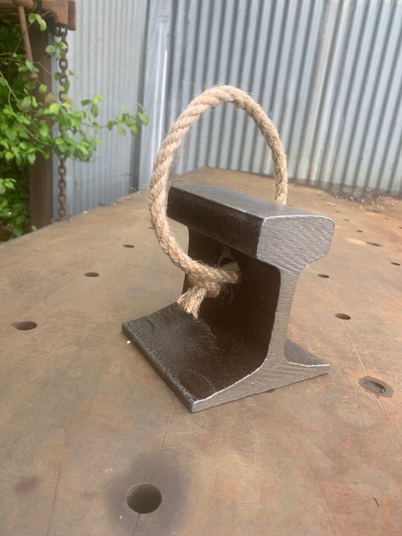 May include: A dark gray, industrial-style paperweight made from a section of railroad track. A loop of thick, natural-colored rope is attached to the top. The paperweight sits on a weathered metal surface, suggesting a rustic or vintage aesthetic.