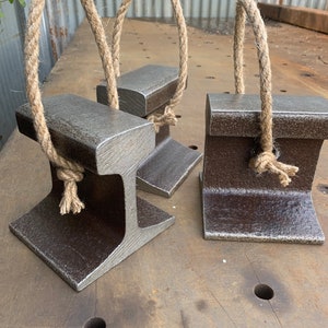 May include: Three metal doorstops with a rustic, industrial design. Each doorstop is a dark gray color and features a sturdy, I-beam shape with a natural rope handle. The doorstops are displayed on a textured metal surface.