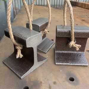 May include: Three metal doorstops with a rustic, industrial design. Each doorstop is a dark gray color and features a sturdy, I-beam shape with a natural rope handle. The doorstops are displayed on a textured metal surface.