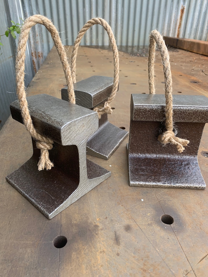 May include: Three metal doorstops with a rustic, industrial design. Each doorstop is a dark gray color and features a sturdy, I-beam shape with a natural rope handle. The doorstops are displayed on a textured metal surface.