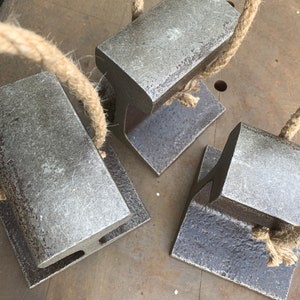 May include: Four weathered metal doorstops with attached rope handles rest on a metal surface. The doorstops are rectangular with a T-shaped base. The rope is a natural tan color.
